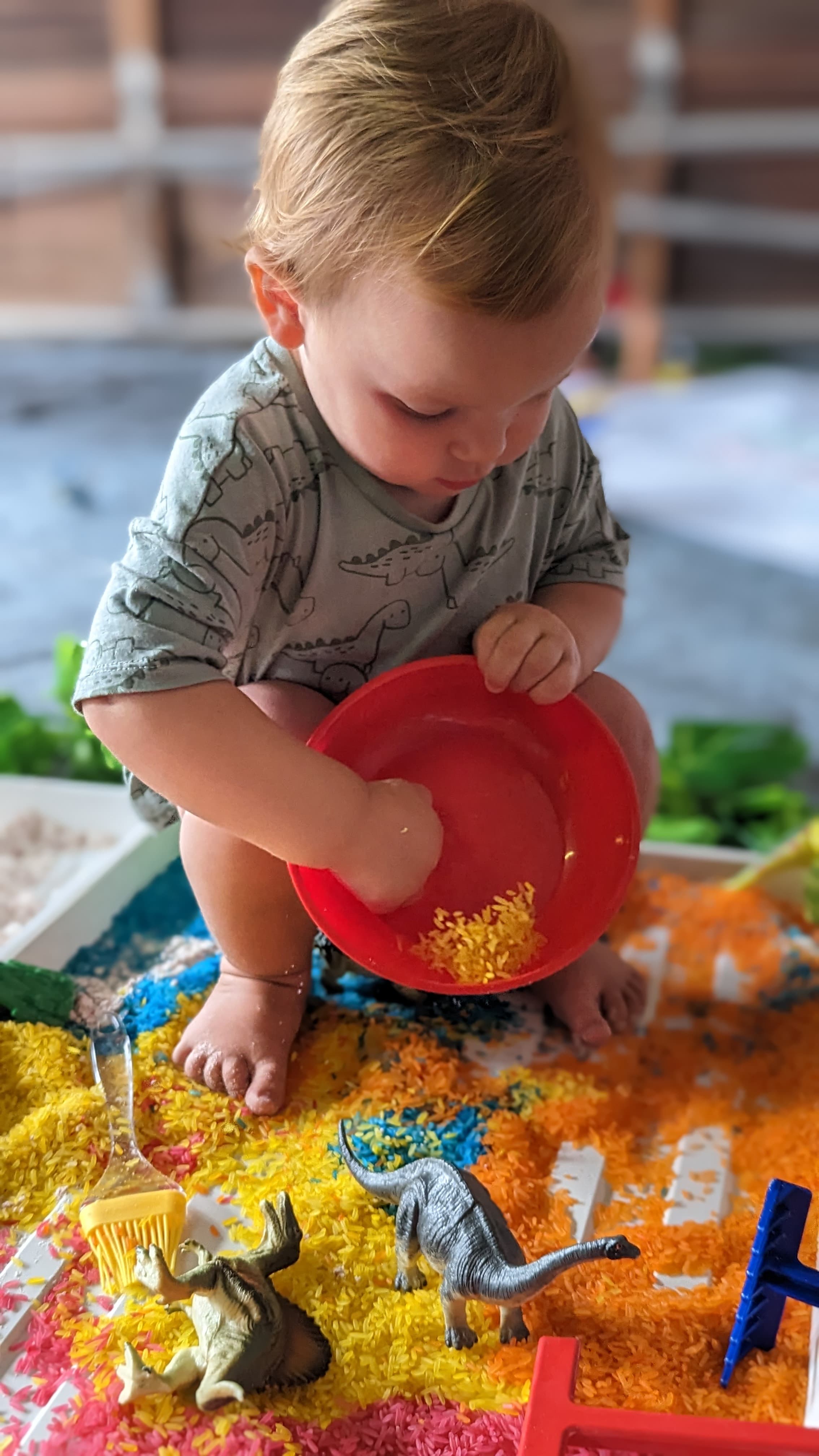 A toddler playing happily in a tray of messy, dinosaur-themed toys and materials.